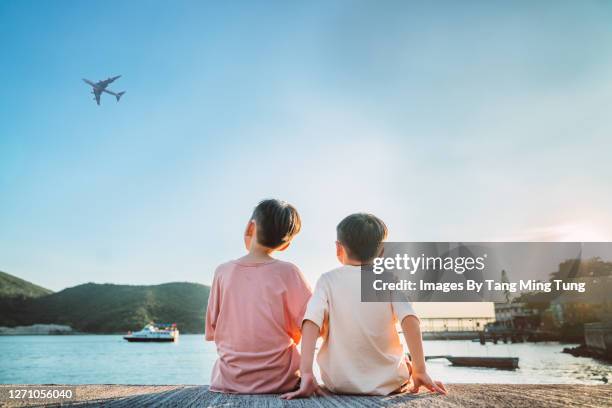 rear view of two little brothers sitting side by side, looking at airplane in pier during sunset. - b��ro stock-fotos und bilder