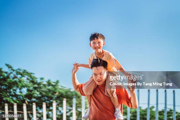cheerful son riding on his dad’s shoulders in park joyfully - asian father son foto e immagini stock