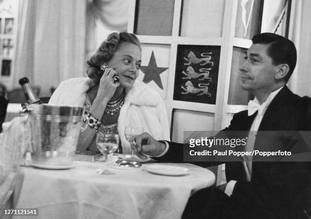 Norwegian former figure skater and film star Sonja Henie , wearing a white fur coat along with diamond and emerald jewellery, drinks champagne with...