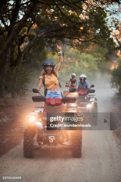 young woman smiling wearing helmet hand in the air riding quad bike being followed by friends in the forest - atv riding stock pictures, royalty-free photos & images