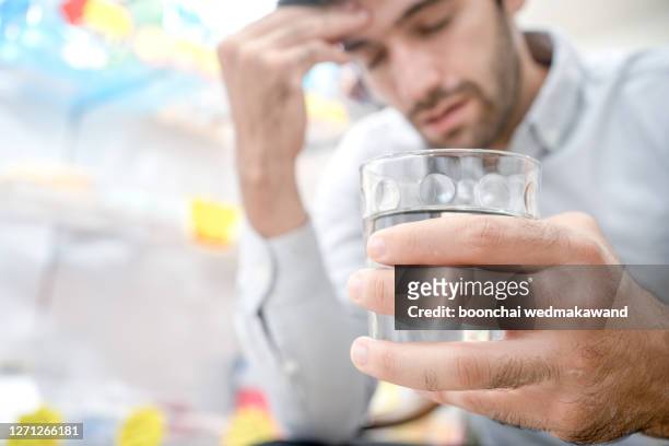 young man suffering from strong headache or migraine sitting with glass of water in the kitchen, millennial guy feeling intoxication and pain touching aching head, morning after hangover concep - chronic fatigue stock-fotos und bilder