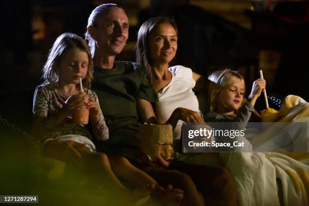Happy family sitting on sofa enjoying movie night outdoor at home backyard together. Mother, father and children watching projector, TV, movies with popcorn in the evening