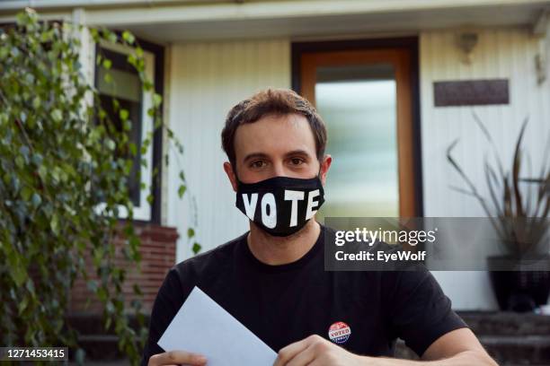 portrait of a man sitting in front of his house, wearing a vote facemask and holding his vote by mail ballot. - voting mask stock pictures, royalty-free photos & images