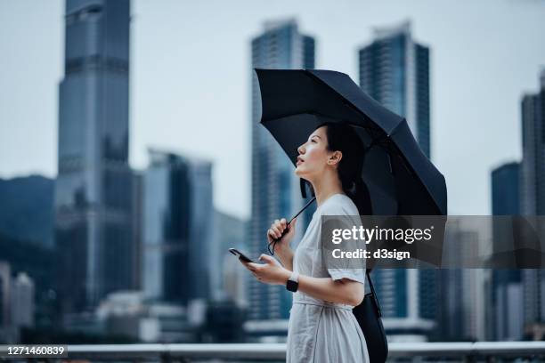 young asian woman looking up to sky feeling the raindrop while walking in the rain in the city street, holding an umbrella and smartphone against urban city scene on a gloomy day. business on the go concept - weather app stock pictures, royalty-free photos & images