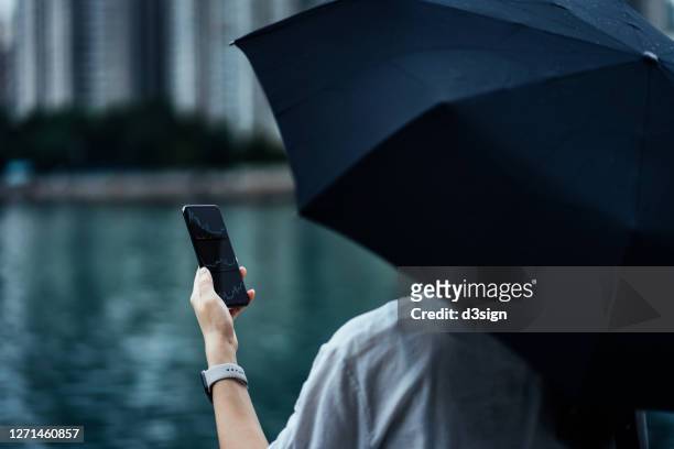 over the shoulder view of young asian businesswoman reading financial trading data on smartphone, holding an umbrella by the promenade in city in the rain, against urban city scene on a gloomy day. business on the go concept - shock stock photos et images de collection