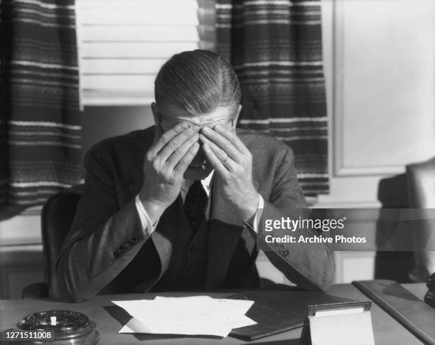 Businessman sits at his office desk, his forehead held in his hands, his elbows propped up on the desk before him, circa 1945. An ashtray and...