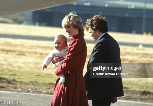 Princess Diana, wearing a burgundy wool coat dress designed by Caroline Charles, carries Prince Wiliiam as she arrives at Aberdeen Airport on March...