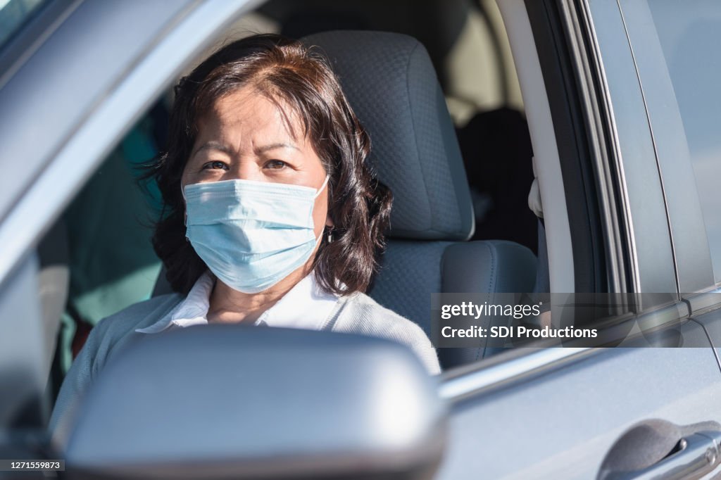Hogere volwassen vrouw, die beschermend masker draagt, kijkt uit autovenster