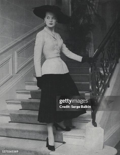 Vogue, April 01, 1947 - Model standing on a staircase wearing a Dior suit comprised of a pale tussore hip-padded jacket, nipped in at the waist, and...
