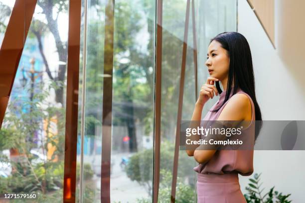 nachdenkliche asiatische geschäftsfrau schaut aus dem fenster in modernes büro - entscheidung stock-fotos und bilder