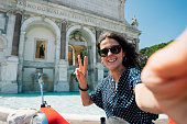 Beautiful young adult woman is taking a selfie on a motor scooter near Fontana dell'Acqua Paola in Rome, Italy