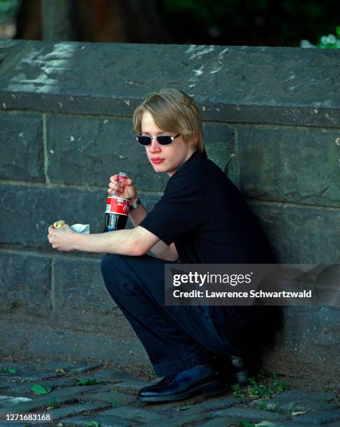 Macaulay Culkin squats on Fifth Avenue near the Metropolitan Museum to eat a hotdog and drink Coca Cola.