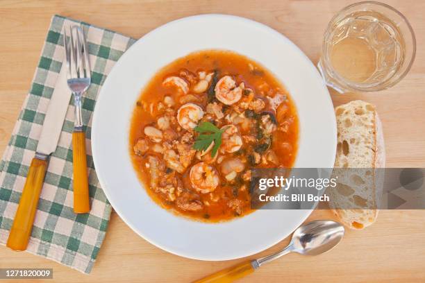 bowl of shrimp gumbo with parsley garnish on a wood table with a glass of white wine, crusty bread, napkin and eating utensils - eating cajun food stock pictures, royalty-free photos & images
