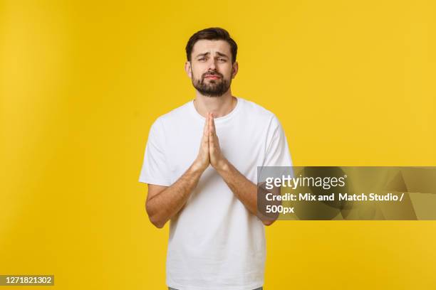 portrait of man standing against yellow background, making praying gesture - rogar fotografías e imágenes de stock