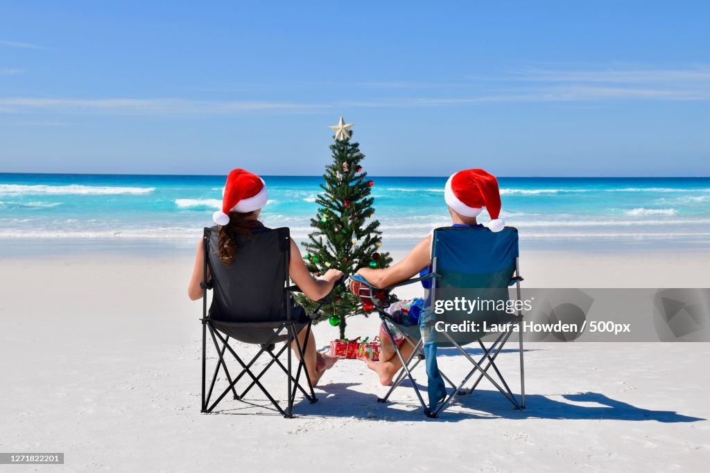 Rear View Of Couple Sitting On Chairs At Beach In Front Of Christmas Tree, West Beach, Australia
