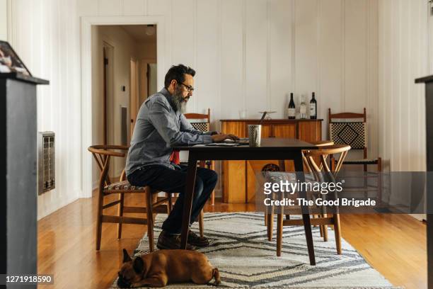 mature man working on laptop in dining room at home - dining table stock pictures, royalty-free photos & images