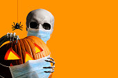 Skeleton Peeks Out From Behind A Jack O'Lantern As Both Wear Protective Face Mask