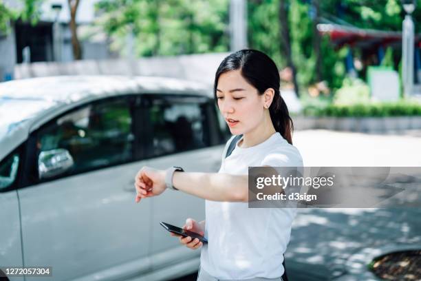 young asian woman being late and in a hurry. she is checking time on smartwatch while waiting for the pick up by the reserved crowdsourced taxi. business on the go concept - chinese car home stock-fotos und bilder