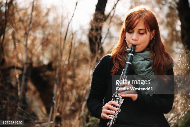 beautiful redhead woman practicing clarinet in forest - klarinette stock-fotos und bilder
