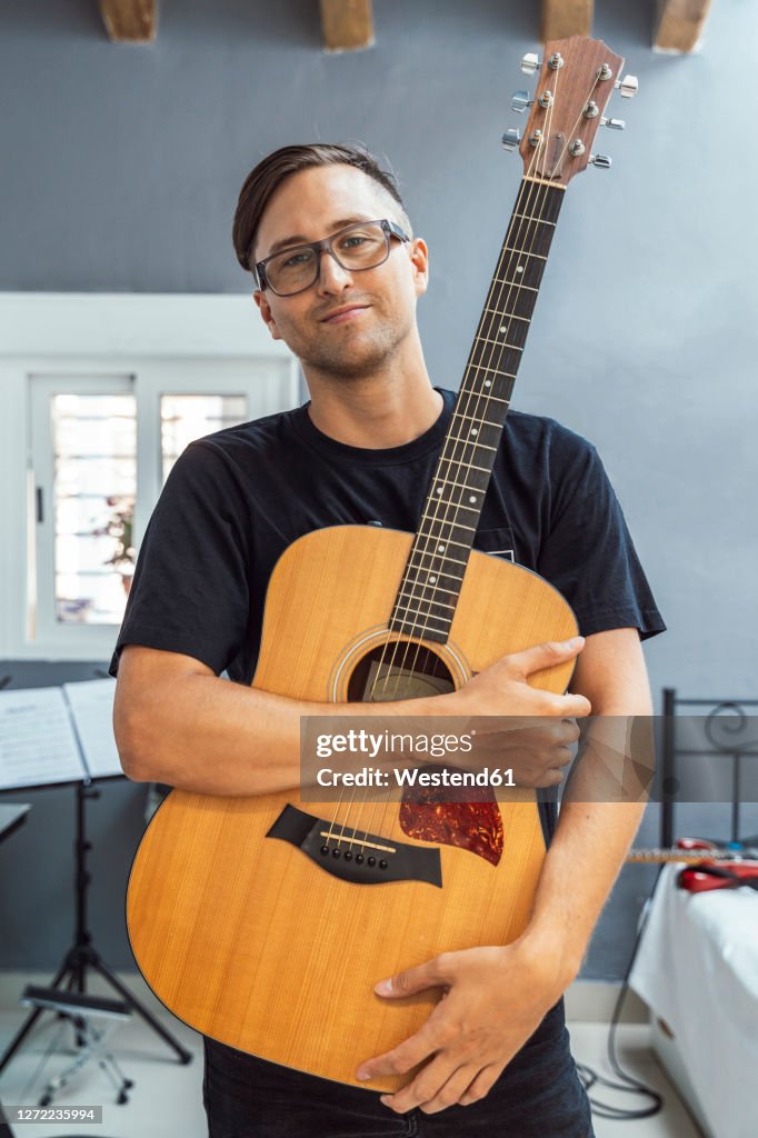 Confident musician holding guitar against wall at home