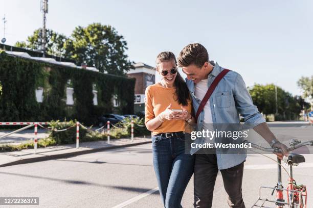 cheerful young couple looking at smartphone while walking on street in city - gedeelde mobiliteit stockfoto's en -beelden