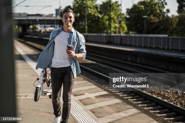 smiling young man carrying push scooter while walking on railroad station platform - man with scooter stock pictures, royalty-free photos & images