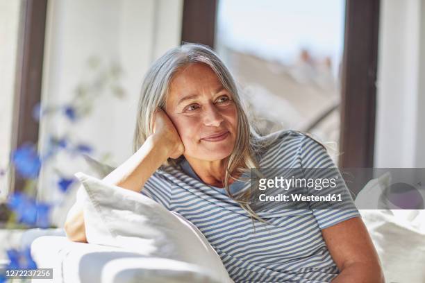 smiling woman relaxing on sofa in living room - cabello gris fotografías e imágenes de stock