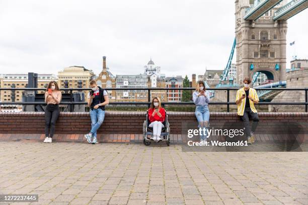 friends maintaining safe distance and using mobile phones with tower bridge in background, london, uk - social distancing friends stock pictures, royalty-free photos & images