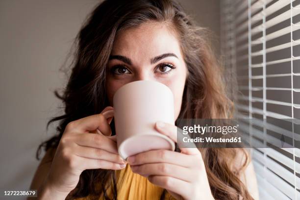 close-up of young woman with long brown hair drinking coffee by blinds at home - cup portraits foto e immagini stock