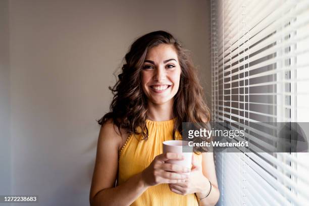 beautiful woman holding coffee cup while standing by window blinds at home - solo una donna giovane foto e immagini stock