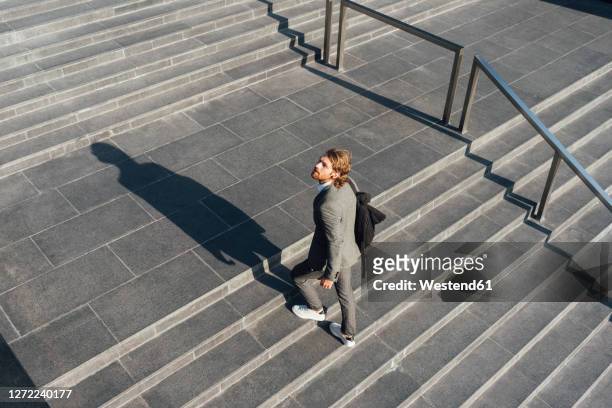male entrepreneur with bag moving up on staircase while looking away in downtown during sunny day - hinauf bewegen stock-fotos und bilder