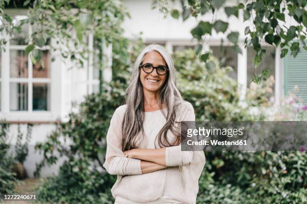 portrait of smiling woman with long grey hair wearing spectacles standing in the garden - frau stock-fotos und bilder
