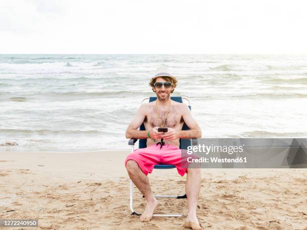 smiling man using smartphone sitting in beach lounger on the beach - badehose stock-fotos und bilder