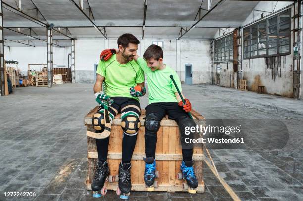 happy boy with arm around father while sitting with hockey sticks on wooden box at court - hockey talks stock pictures, royalty-free photos & images