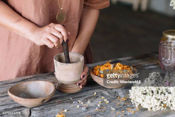 midsection of woman crushing flowers and herbs in mortar with pestle on wooden table - earthenware stock pictures, royalty-free photos & images