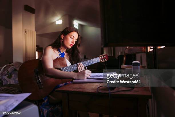 young woman writing in book while practicing guitar at home - zanger en componist stockfoto's en -beelden