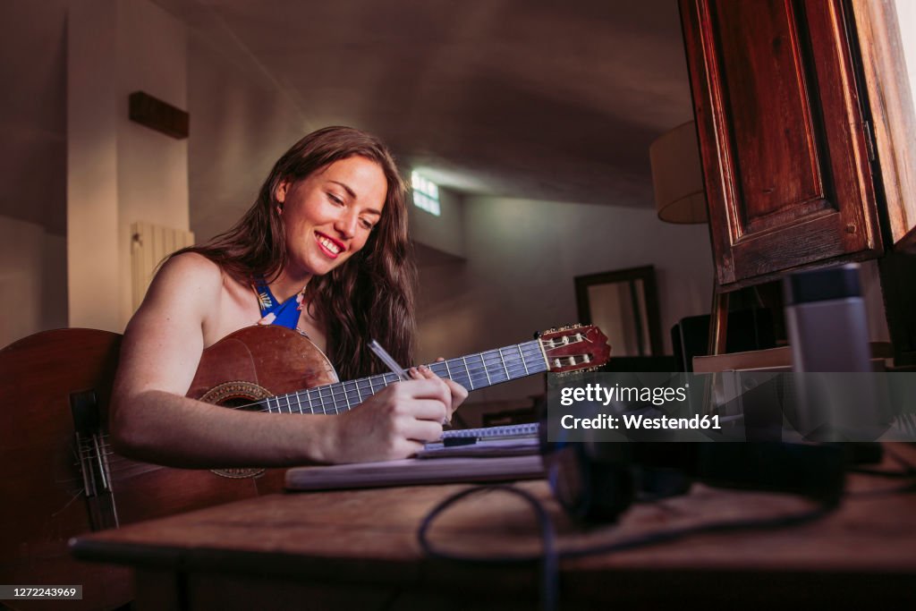 Smiling young woman writing in book while practicing guitar at home