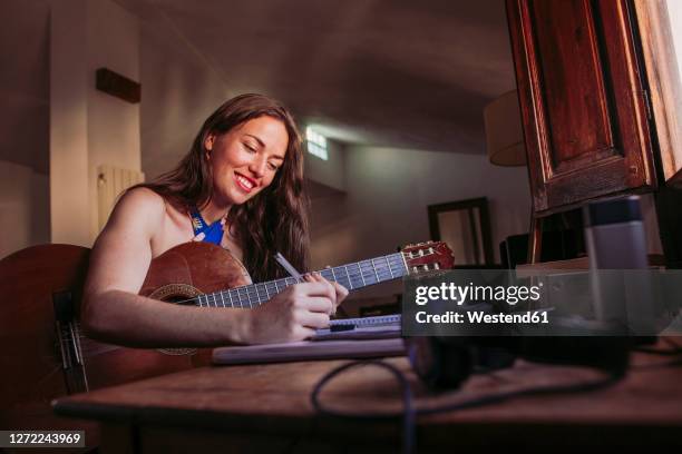 smiling young woman writing in book while practicing guitar at home - singer songwriter stock pictures, royalty-free photos & images