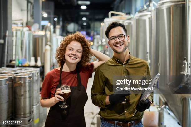 portrait of happy man and woman holding clipboard and beer glass in craft brewery - clipboard and glasses imagens e fotografias de stock