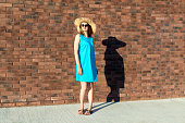Slender Caucasian woman in blue dress and straw hat with fatty shadow on the wall. The concept of inadequate perception of one's own weight. Slimming or fatness symbol