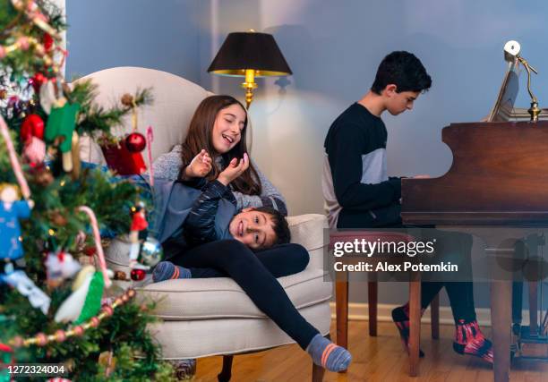 10-years-old girl, a sister, plating with her 5-years-old brother near by the decorated christmas tree when their older brother playing piano in the backdrop. - celebrating 15 years stock pictures, royalty-free photos & images