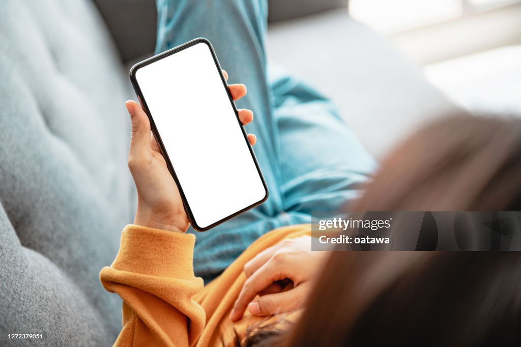 Woman using mobile smartphone with blank white screen on a sofa in living room.