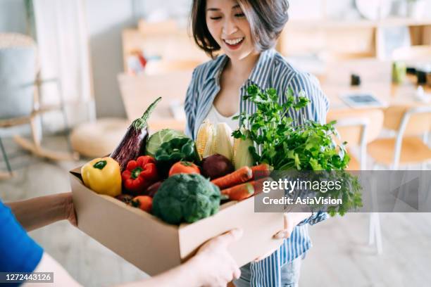 cheerful young asian woman receiving a box full of colourful and fresh organic groceries ordered online from delivery person at home - grocery food stock pictures, royalty-free photos & images