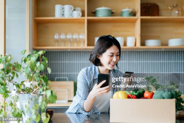 beautiful smiling young asian woman grocery shopping online with mobile app device on smartphone and making online payment with her credit card, with a box of colourful and fresh organic groceries on the kitchen counter at home - asia food stock pictures, royalty-free photos & images