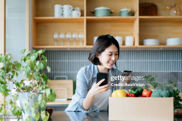 beautiful smiling young asian woman grocery shopping online with mobile app device on smartphone and making online payment with her credit card, with a box of colourful and fresh organic groceries on the kitchen counter at home - e commerce payment stock pictures, royalty-free photos & images