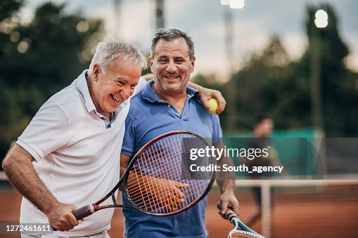 Two cheerful senior men talking while walking on the outdoor tennis court Two cheerful senior men talking while walking on the outdoor tennis court