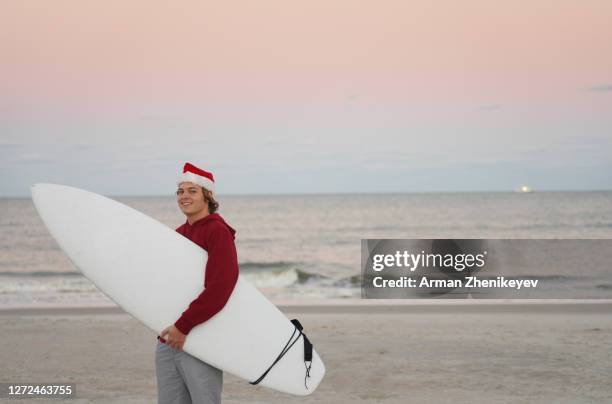 man dressed as santa claus carrying surfboard next to the ocean - surfing santa stock pictures, royalty-free photos & images