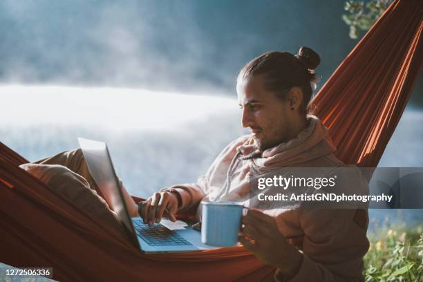 man in hammock using laptop, drinking coffee and relaxing around lake at sunrise. - development camp stock pictures, royalty-free photos & images