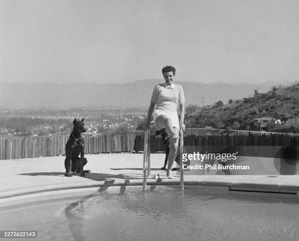 American actress Jane Russell leans on the ladder of a pool as she lifts one leg up with her two Doberman dogs, Blitz and Katherine, by her side, US,...