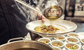 Chef pours broth into a chicken soup with noodles, meat and vegetable.